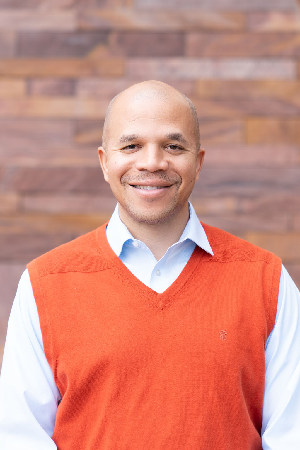 man smiling with orange sweater on surrounded by brick wall